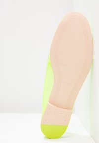 Semelle d'une chaussure plate jaune fluo avec un dessous et un talon beiges lisses, marquée "ALDO," posée en angle contre un mur blanc.