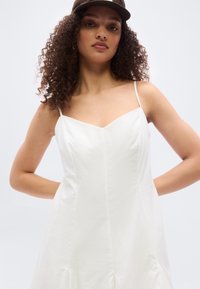 Νεαρή γυναίκα με σγουρά μαλλιά, φορώντας λευκό αμάνικο φόρεμα και μαύρο καπέλο, ποζάρει με τα χέρια πίσω από την πλάτη της μπροστά σε λευκό φόντο.