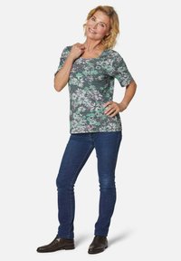 Bloemmotief korte mouwen top in groen, roze en wit op een donkere achtergrond, gecombineerd met strakke blauwe jeans en donkere enkellaarsjes.