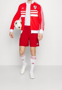 Chaqueta atlética roja con rayas y logo blancos, pantalones cortos rojos, calcetas blancas con rayas rojas y botas de fútbol ligeras. Sostiene un balón de fútbol.