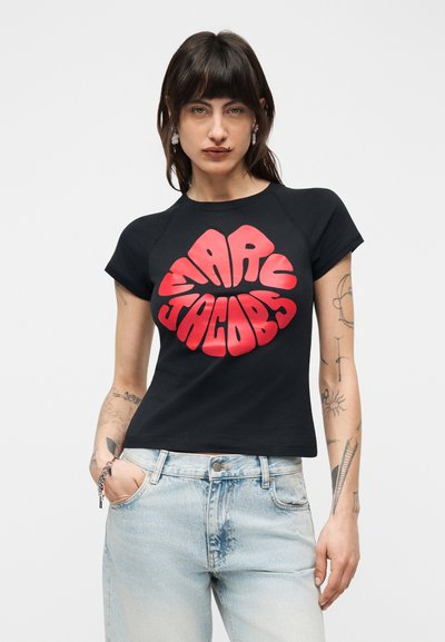 Γυναίκα με σκούρα μαλλιά που φοράει μαύρο crop top με κόκκινο κείμενο «Marc Jacobs» και ανοιχτόχρωμα τζιν ψηλόμεσα, ποζάρει με το χέρι στην τσέπη.