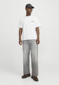 Camiseta blanca con un pequeño logo de color, combinada con jeans de pierna ancha gris claro y botas marrones. Gorra negra con acentos de texto.