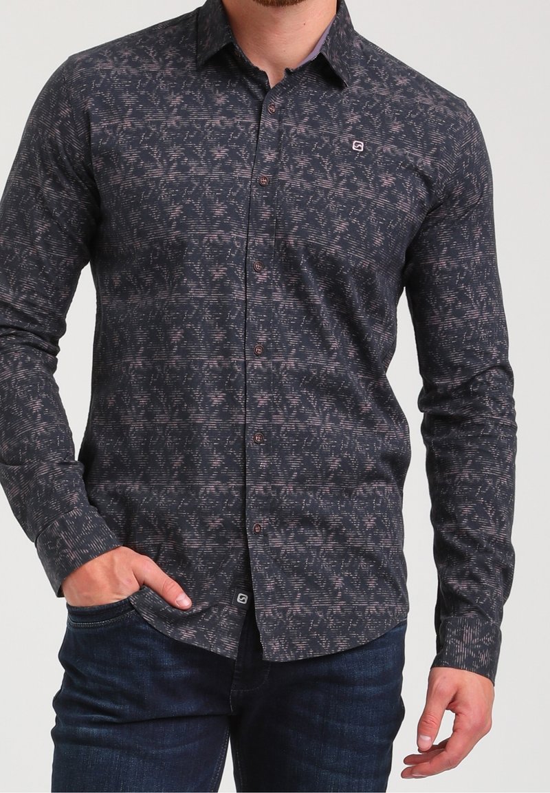 Homme portant une chemise boutonnée sombre avec un motif abstrait subtil rose et gris et un jean bleu foncé, main dans la poche.