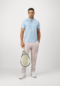 Lys blå polo-skjorte med en logo, paret med lyse rosa treningsbukser. Holder en tennisracket, iført hvite sko med svarte detaljer.