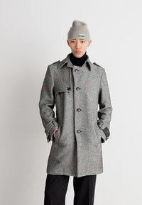 Manteau à chevrons gris avec de grands boutons, épaulettes et accents en cuir sur les poignets. Porté par-dessus un col roulé noir et un pantalon noir.