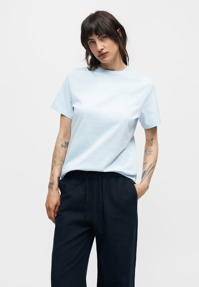 Άτομο με σκούρα μαλλιά μέχρι τον ώμο και τατουάζ στα χέρια, φοράει ένα ανοιχτό μπλε κοντομάνικο πουκάμισο και μαύρο παντελόνι, στέκεται με το ένα χέρι στην τσέπη.