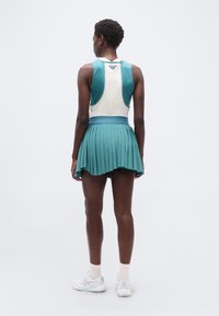 Γυναίκα που στέκεται με την πλάτη γυρισμένη στην κάμερα, φο ράει τιρκουάζ και λευκό μπλουζάκι χωρίς μανίκια, τιρκουάζ φούστα με πτυχώσεις, λευκές κάλτσες και λευκά αθλητικά παπούτσια.