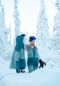 Snow gear set includes a teal jacket and matching jumpsuit with light blue accents, both featuring zippers and pockets, in a snowy forest setting.