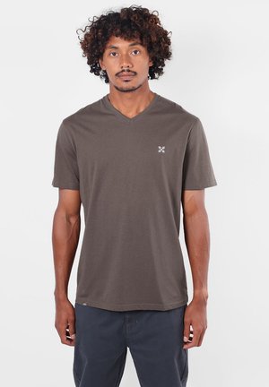 Homme aux cheveux bouclés portant un t-shirt marron uni à col en V et un pantalon foncé, debout devant un fond blanc.