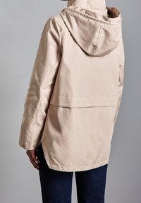 Ljusbeige vattentät jacka med huva, med en avslappnad passform, utan synliga knappar och detalj med ventilerad fåll. Slät textur.