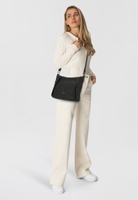 Schwarze, strukturierte Handtasche mit Textur und verstellbarem Träger. Das Model trägt ein helles, geripptes Oberteil und weite Hosen.