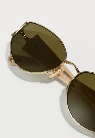 Gafas de sol en tono dorado con grandes lentes ovalados de color verde oscuro y un marco ligero de color beige. Presentan un diseño minimalista y una marca sutil.