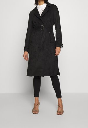 Vrouw draagt een zwarte getailleerde trenchcoat, zwarte leggings, witte gebreide top en sandalen met hoge hakken en bandjes, staand tegen een effen achtergrond.