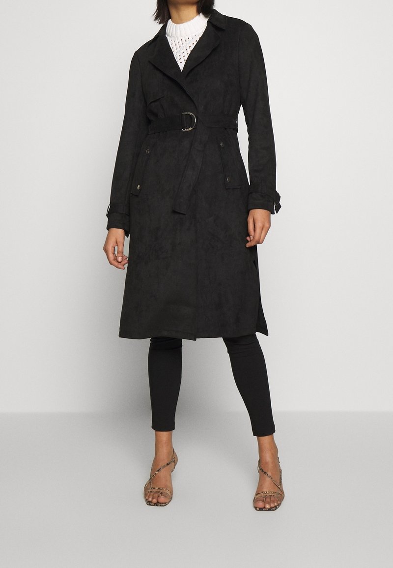 Femme portant un trench-coat noir ceinturé, des leggings noirs, un top en maille blanc et des sandales à talons hauts à brides, debout devant un fond uni.