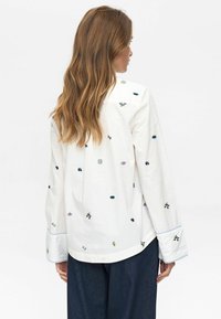 Vrouw met lang bruin haar draagt een witte blouse met kleine kleurrijke geborduurde ontwerpen, in combinatie met donkerblauwe wijde broek, met haar rug naar ons gekeerd.