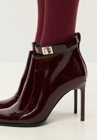 Bottines en cuir verni bordeaux à talon haut, avec une sangle à boucle autour de la cheville et une finition lisse et brillante.