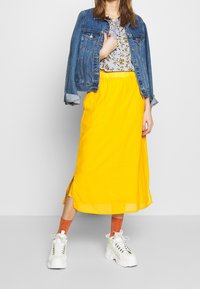 Giacca di jeans, blouse floreale e gonna midi gialla con spacco laterale. Indossata con sneakers bianche chunky e calzini arancioni.