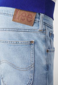 Jeans de mezclilla azul claro con un parche de cuero de la marca "Lee", con detalles de costura visibles y un corte ajustado.