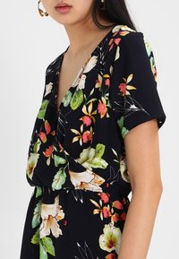 Kvinna i en svart blommig omlottskarka med korta ärmar och färgglada gröna, röda, gula och vita blommor på vit bakgrund.