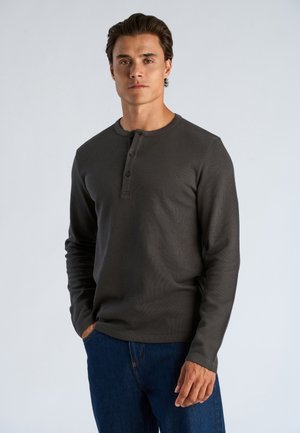Man draagt een donkergrijs henley shirt met lange mouwen en een blauwe spijkerbroek, staand met één hand in de zak voor een egale achtergrond.