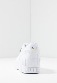 Sneaker bianche con tomaia in pelle texture, forma arrotondata, suola bianca spessa e dettaglio del logo dorato sul tallone. Presentano un accento in nastro.