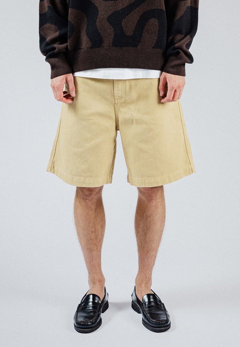 Homme portant un short beige longueur genou, des mocassins noirs et un pull foncé à motifs, debout devant un fond uni.