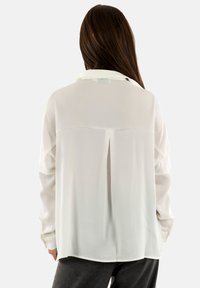 Blusa blanca de manga larga con cuello, con un respaldo plisado, corte suelto y tejido texturizado. El modelo la combina con pantalones grises.