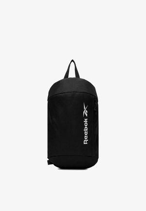 Mochila tipo bandolera negra de Reebok con logo vertical blanco, compartimento principal con cremallera y asa superior para transportar, sobre fondo blanco.