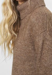 Pull à col roulé en tricot marron avec une texture douce, une coupe décontractée et un col à côtes pour plus de chaleur. Détail marbré notable dans le fil.
