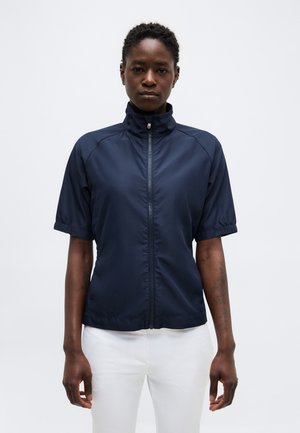 Persoon met kort krullend haar, die een marineblauw kortemouws jack met rits en witte broek draagt, staand tegen een effen achtergrond.