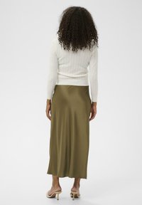 Femme avec des cheveux bouclés portant un pull en tricot blanc, une jupe midi vert olive et des sandales à talons beiges, se tenant de dos sur fond blanc.