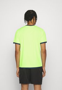 Hombre con cabello trenzado de espaldas, vistiendo una camiseta de neón verde brillante con ribetes oscuros y pantalones cortos negros frente a un fondo liso.