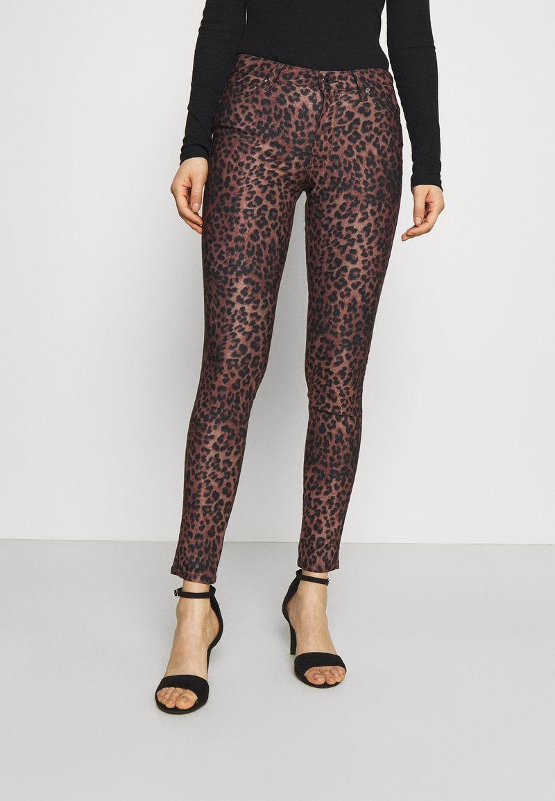 Femme portant un pantalon slim marron et noir à imprimé léopard, un haut noir à manches longues et des sandales noires à talons hauts, debout sur un sol clair.
