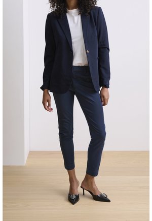 Femme debout dans un blazer marine, un pantalon slim assorti, une chemise blanche et des escarpins noirs à bout pointu sur un sol en bois clair contre un mur blanc.