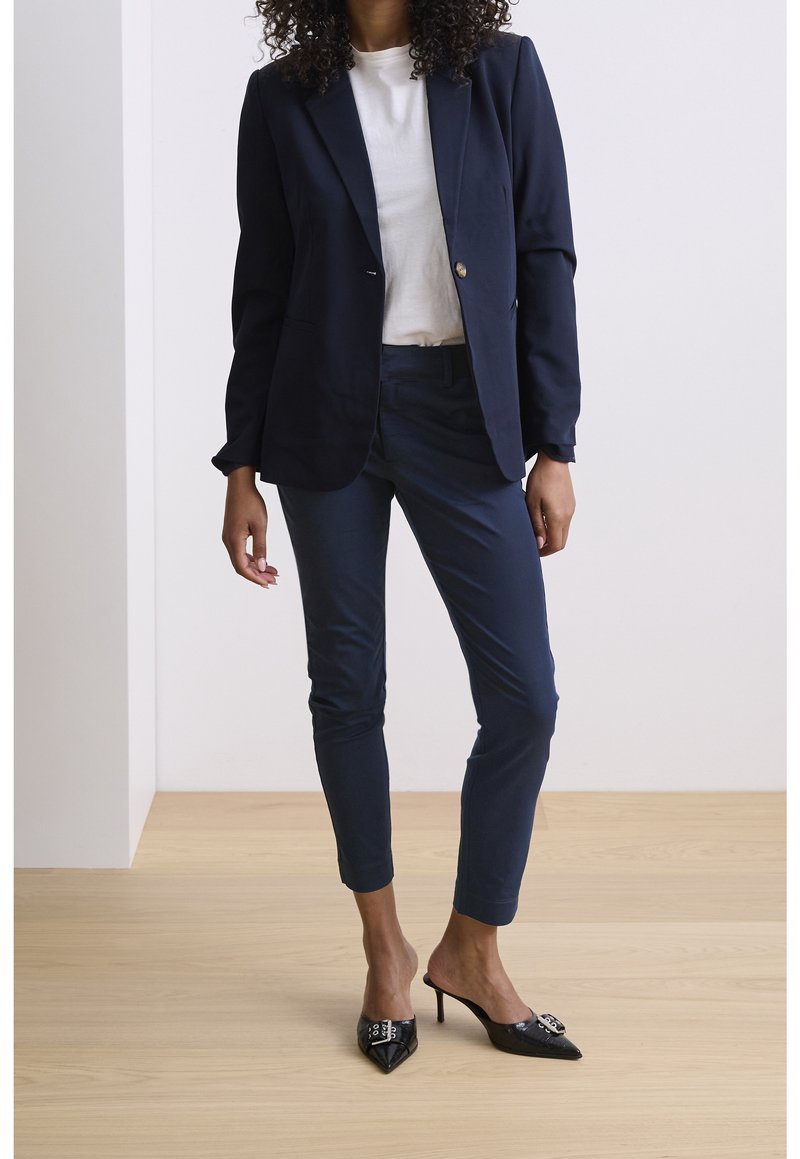 Femme debout dans un blazer marine, un pantalon slim assorti, une chemise blanche et des escarpins noirs à bout pointu sur un sol en bois clair contre un mur blanc.