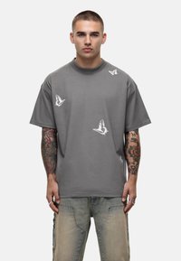 Grijze katoenen t-shirt met korte mouwen, voorzien van witte vlinderprints verspreid op de voorkant. Ronde hals met een relaxte pasvorm.