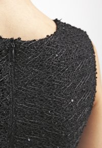 Tissu noir texturé avec un motif en relief, comprenant de petites embellissements. Fermeture éclair arrière visible au niveau du cou.