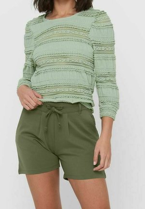 Femme portant un haut texturé à manches longues vert clair et un short à cordon vert olive sur un fond clair uni.