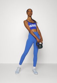 Zila sporta krūšturis ar baltu uzrakstu, atbilstošas zilas augsta jostasvietas legingas un gaiši zilas sporta kurpes, tur rokā melnu kettlebellu.