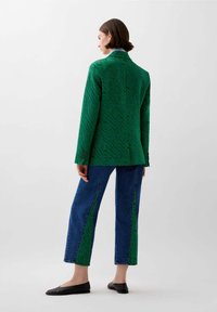 Blazer verde con texture a zigzag, abbinato a pantaloni in denim blu con pannelli laterali verdi. Scarpe flat nere completano il look.