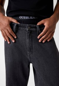 Zwarte denim jeans met een rechte pijp, voorzien van gestikte details, een metalen knoop en een elastische tailleband met de tekst "GUESS JEANS".