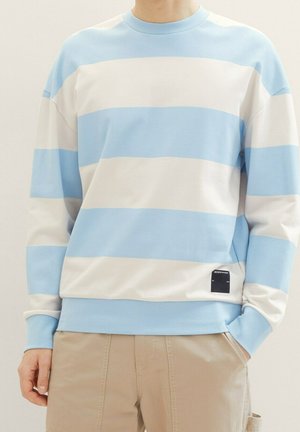 Mann trägt beige Hosen und einen Pullover mit langen Ärmeln, der breite horizontale hellblaue und weiße Streifen hat, die Hände locker an den Seiten.