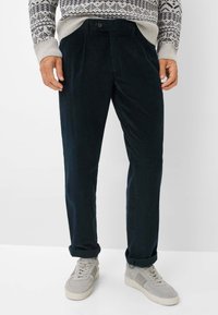 Uomo che indossa pantaloni in velluto a coste blu scuro arrotolati alle caviglie, sneakers grigio chiaro e un maglione a motivi con design a fiocchi di neve.