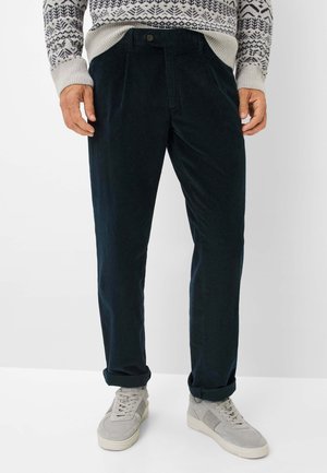 Mann in dunkelblauen Cordhosen mit hochgerollten Säumen, hellgrauen Turnschuhen und einem gemusterten Pullover mit Schneeflockenmotiven.