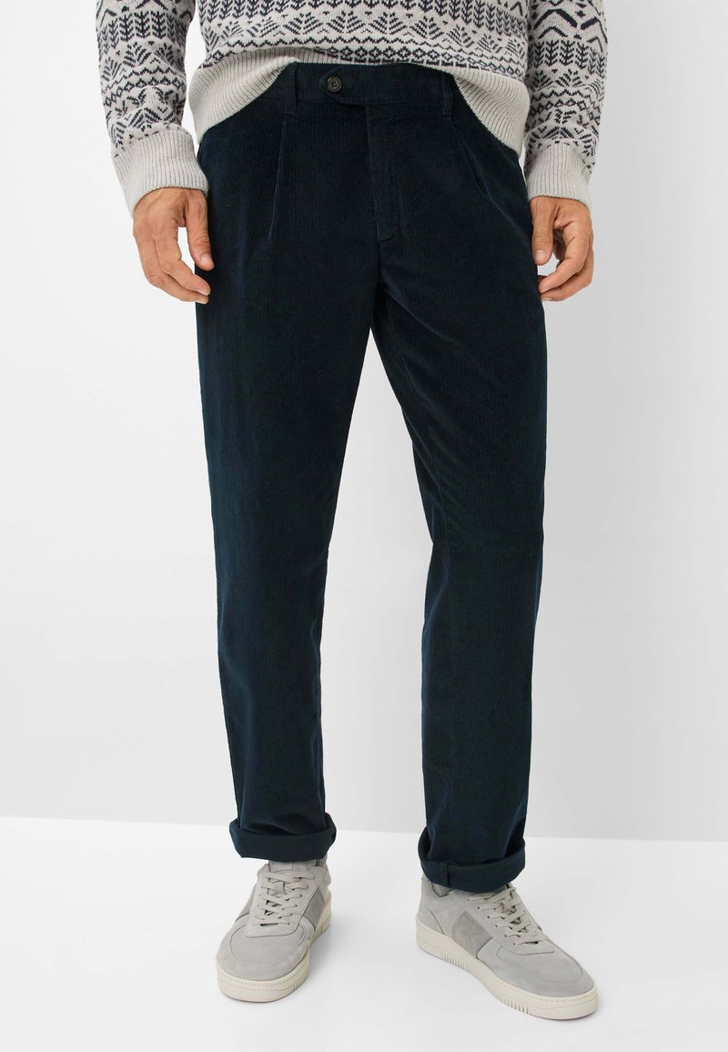 Uomo che indossa pantaloni in velluto a coste blu scuro arrotolati alle caviglie, sneakers grigio chiaro e un maglione a motivi con design a fiocchi di neve.