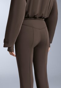 Hnedé legíny s priliehavým strihom a hladkou textúrou, s vysokým pásom a viditeľnými švovými detailmi. Materiál vyzerá elasticky a dobre sa prispôsobuje postave.