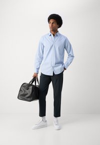 Lyseblå rutete skjorte med knapper, mørke bukser, hvite sneakers, holder en svart teksturert treningsveske. Minimalistisk og moderne design.