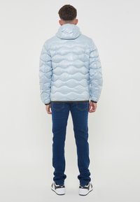 Giubbotto puffer azzurro chiaro con finitura lucida, modello a onde trapuntato, cappuccio regolabile e polsini neri, abbinato a jeans di denim scuro e sneaker.