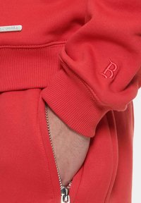Roter Sweatshirt mit Rippbündchen, einer Reißverschlusstasche und einem gestickten 'B'-Logo. Das Material wirkt weich und hat eine glatte Textur.