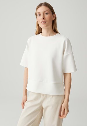 Vrouw met een lichte huid en blond haar, gekleed in een losvallend wit T-shirt met korte mouwen en beige broek, staand tegen een effen lichtgrijze achtergrond.